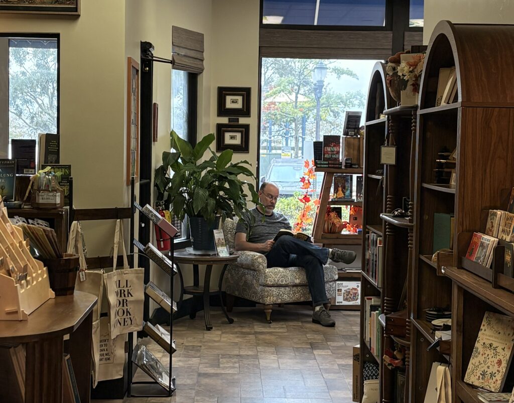 A reader seated in an armchair beside bookshelves and windows at Cashmere Books & Gifts.